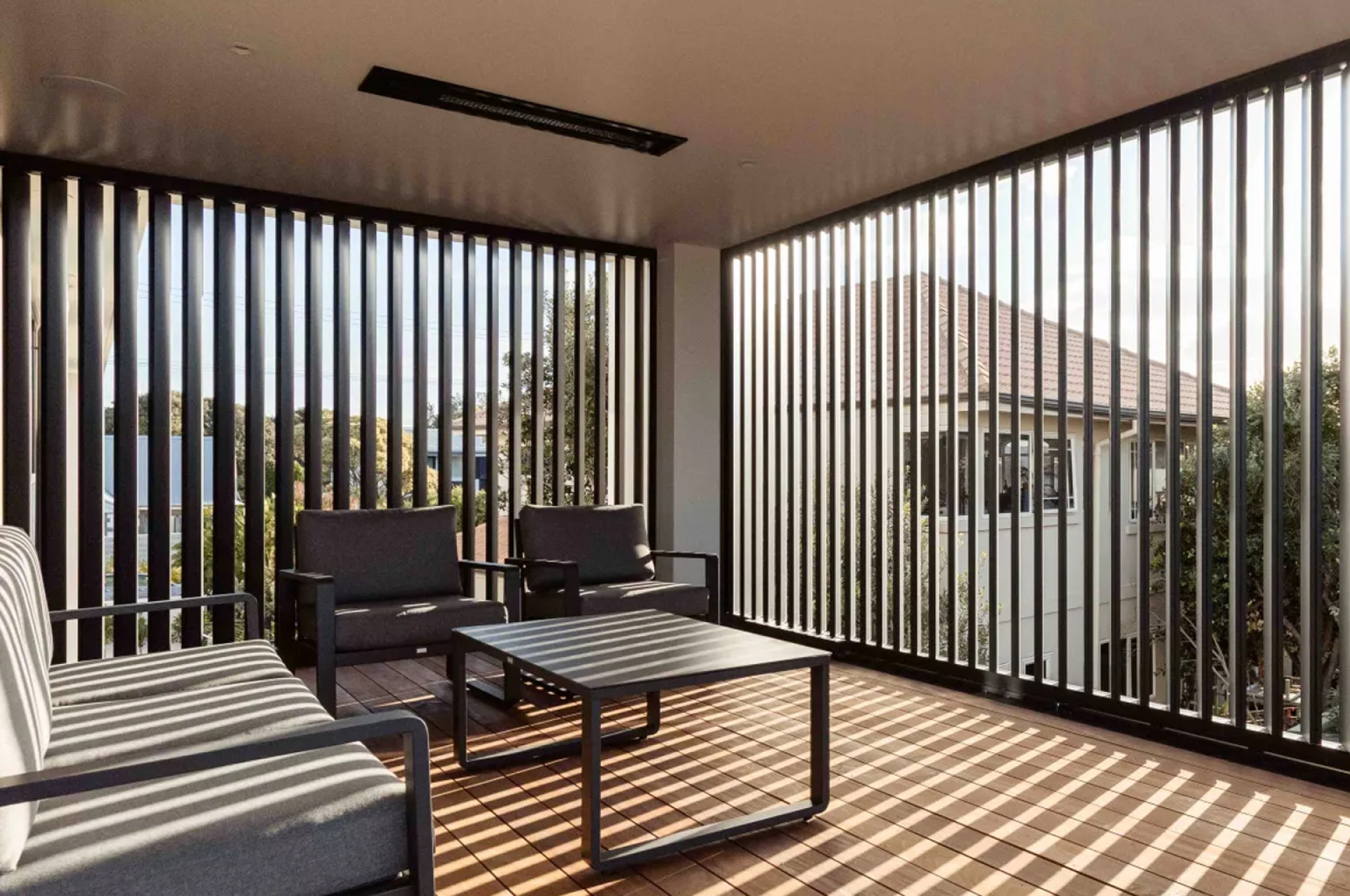 A modern patio with dark furniture and a slatted wall, creating sun stripes on the wooden deck.