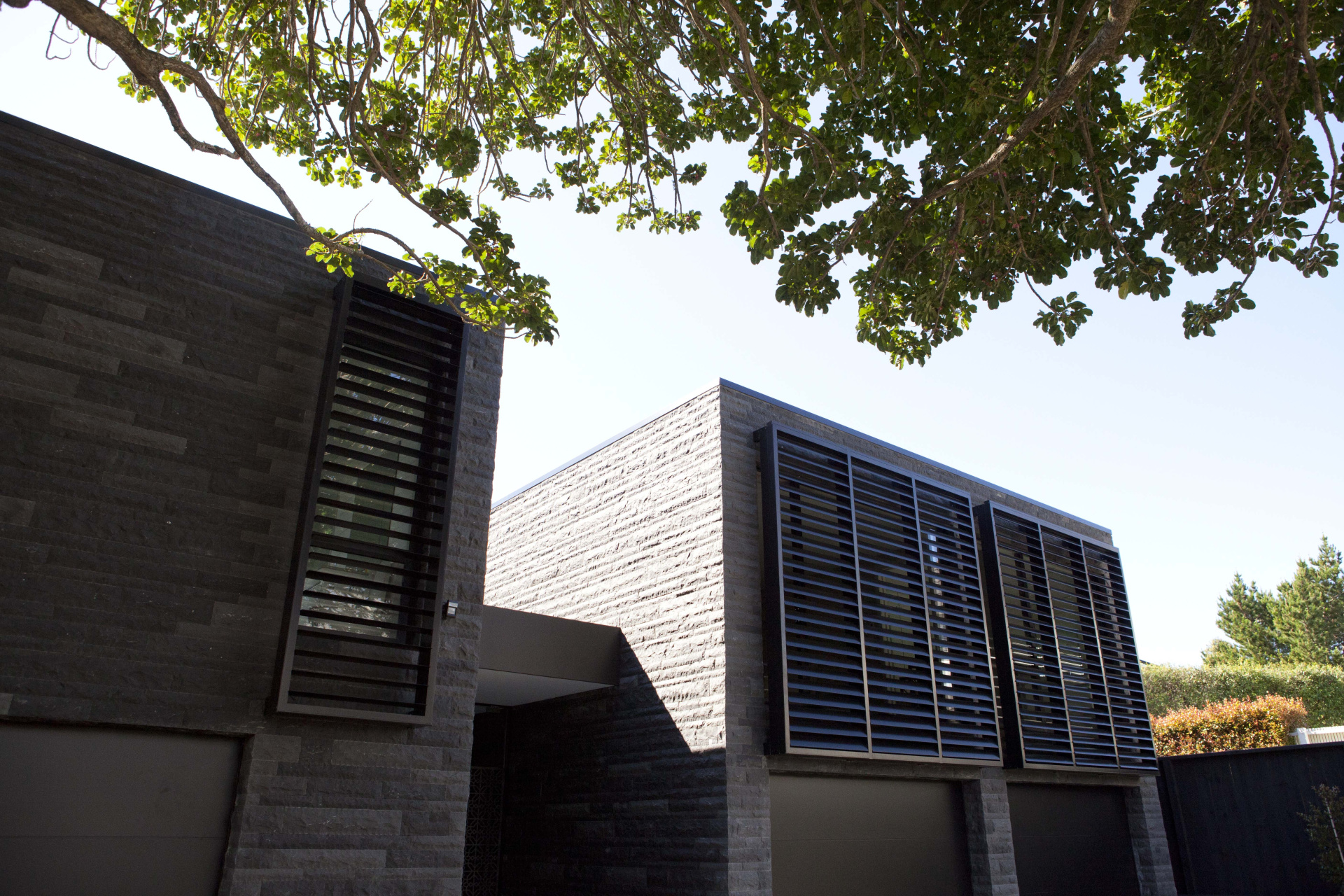 Modern house exterior with dark stone walls, large louvred windows, and tree branches providing shade in the foreground.