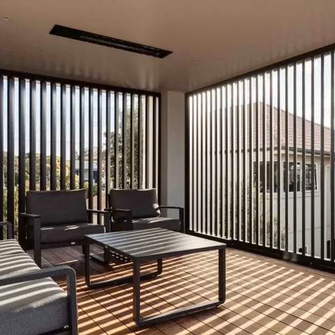 A modern patio with dark furniture and a slatted wall, creating sun stripes on the wooden deck.