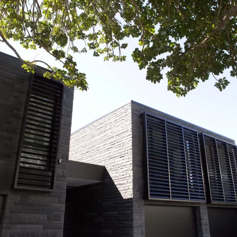 Modern house exterior with dark stone walls, large louvred windows, and tree branches providing shade in the foreground.