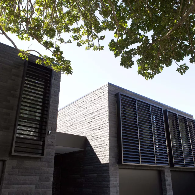 Modern house exterior with dark stone walls, large louvred windows, and tree branches providing shade in the foreground.